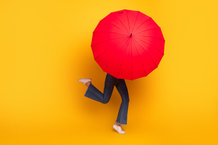 Playful woman concealing herself behind a vibrant red umbrella on a bright yellow backgroundの写真素材