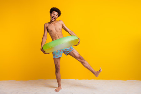 Cheerful shirtless young man with afro hairstyle holding inflatable ring against a vibrant yellow backgroundの写真素材