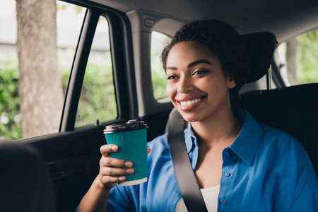Smiling young woman enjoying a coffee while riding in a car on a sunny day in the city, dressed in...