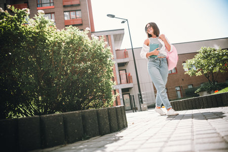 Young woman enjoying a sunny day while walking through an urban street with a book and a stylish outfitの写真素材