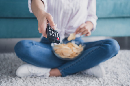 Cropped portrait of pretty young girl hold remote control watch tv eat chips white interior modern...