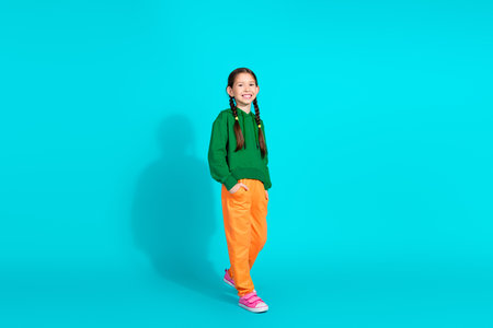 Smiling girl in stylish green hoodie and orange pants posing on a turquoise background.の写真素材