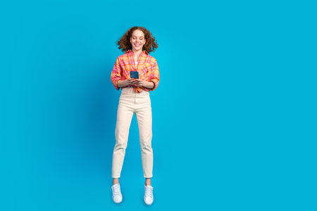 Happy young woman with curly hair using smartphone against blue background, wearing casual outfit,...