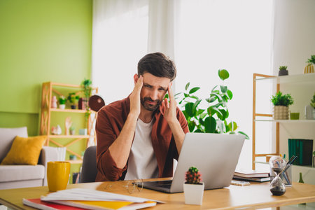 Young bearded man working on a laptop in a modern home office, looking stressed while in a bright...