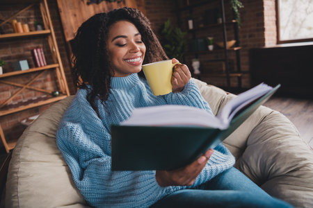 Young woman in a cozy sweater enjoying a book and hot drink in a bright living room during fallの写真素材