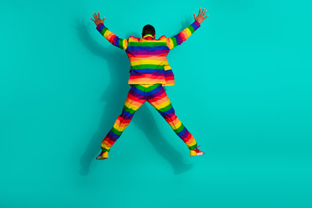 Young man in colorful rainbow striped suit leaping against vibrant turquoise background, expressing personality, diversity, and celebratory joyの写真素材