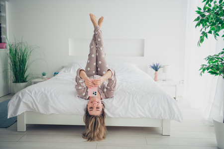 Young woman relaxing on cozy bed in pajamas, using smartphone in natural daylightの写真素材