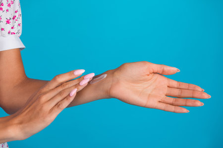 Young womans hands applying moisturizer on a blue background, showcasing skincare routine and...