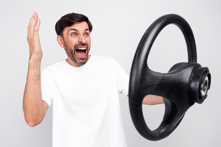 Excited young man posing with his hands up and a steering wheel against a white backgroundの写真素材