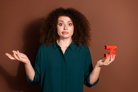 Photo of unsure woman wear formal shirt shrug shoulders holding binoculars isolated brown color backgroundの写真素材