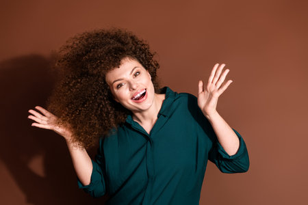 Joyful young woman with curly hair expressing excitement against a brown background, wearing a casual green shirt and smiling brightlyの写真素材
