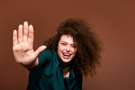 Young woman with curly brunette hair in green shirt smiling against a brown background, radiating positive energy and playfulnessの写真素材