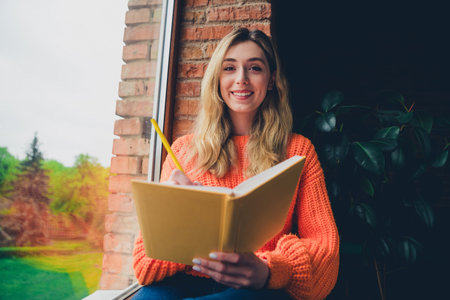 Photo of attractive young woman sit windowsill write diary dressed orange outfit cozy day light home interior living roomの写真素材