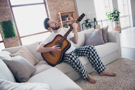 Photo of carefree positive young man in pajama sitting on sofa playing guitar party friday weekend indoorsの写真素材