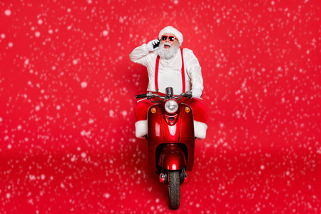 Portrait of nice attractive bearded cheerful funny Santa wearing cap hat riding motor bike talking on phone taking orders isolated over bright vivid shine vibrant red color backgroundの写真素材
