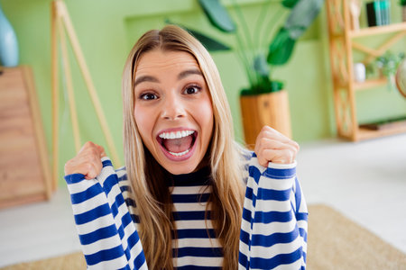 Photo of attractive blonde young woman excited winning raise fists dressed striped outfit cozy day light home interior living roomの写真素材