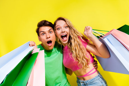 Portrait of two nice young partners open mouth hold mall bags wear t-shirt isolated on vibrant yellow color backgroundの写真素材