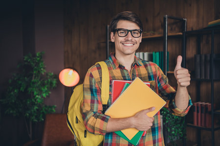 Photo of clever intelligent nice young man wear checkered shirt hold book thumb up indoors collegeの写真素材