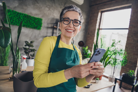 Photo portrait of lovely pensioner lady hold device florist businesswoman wear green apron working flower shop studio small businessの写真素材