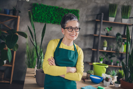 Photo of nice elderly lady crossed hands look empty space wear green apron enjoying gardering indoors home business workplaceの写真素材