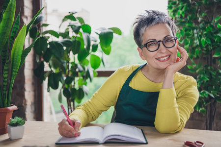 Photo of lovely pensioner lady write notebok planning florist businesswoman wear green apron working flower shop studio small businessの写真素材