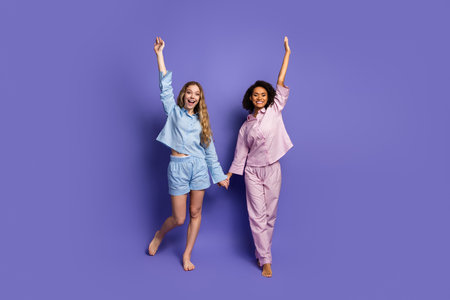 Two young women in comfortable sleepwear expressing joy and friendship against a purple backgroundの写真素材