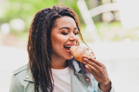Young woman enjoying a delicious croissant outdoors on a sunny day in the city, wearing a stylish jacket during her urban exploration.の写真素材