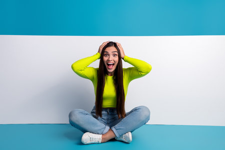 Excited young woman sitting cross-legged against a vibrant blue backdrop, wearing a bright yellow top and expressing joyの写真素材