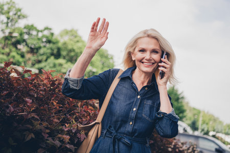 Charming senior woman in denim dress enjoying a sunny day outdoors while talking on phone and waving happilyの写真素材