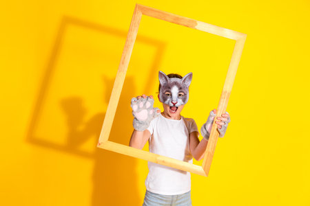 Young girl in a cat mask posing with a wooden frame against a vibrant yellow backgroundの写真素材