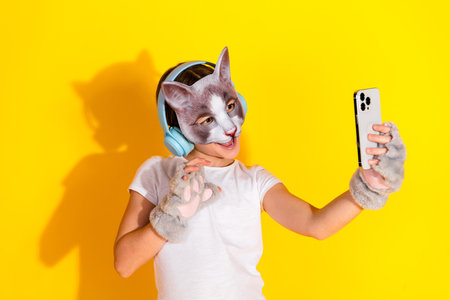 Young girl in a playful cat mask using a phone with a vibrant yellow background exuding cheerful and creative vibes.の写真素材