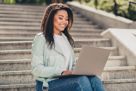 Young woman enjoying daylight on urban steps with a smile, wearing a stylish jacket while working on her laptop outdoors during a warm day.の写真素材