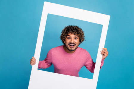 Cheerful young man in a casual pink pullover holding a white frame against a blue background,...