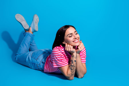 Smiling woman lying on a blue background wearing a pink striped shirt and jeans, presenting a...