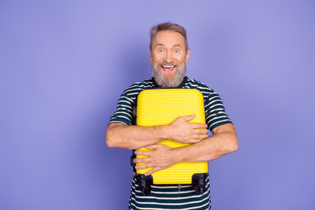 Cheerful elderly man embracing a yellow suitcase against a vibrant purple background, showcasing a trendy lifestyleの写真素材