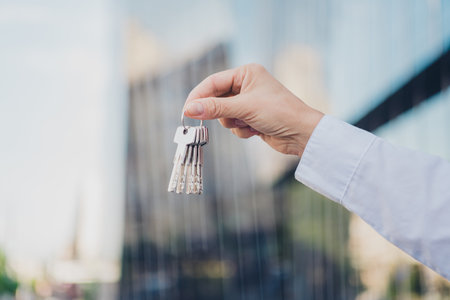 Close-up of a hand holding a set of keys against a modern urban skyscraper background in daylightの写真素材