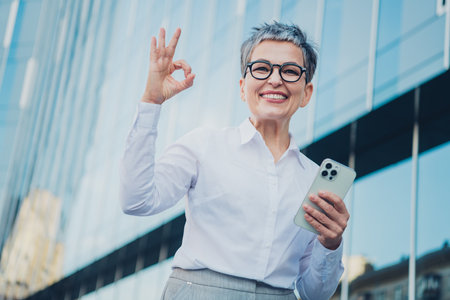 Confident mature woman with short grey hair in formalwear, gesturing OK symbol and holding smartphone, standing outdoors in urban cityscapeの写真素材