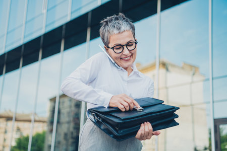 Confident mature businesswoman in the city holding portfolio and talking on phone with a smile, wearing a white shirt and glasses outdoors.の写真素材