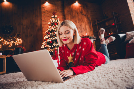 Full length photo of young blonde female lying floor eshopping netbook wear christmas sweater tradition atmosphere decor house indoors roomの写真素材