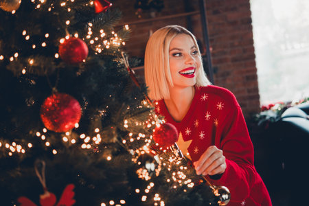 Photo of pretty young blonde female adorn tree wondered dressed christmas sweater tradition atmosphere decor house indoors roomの写真素材