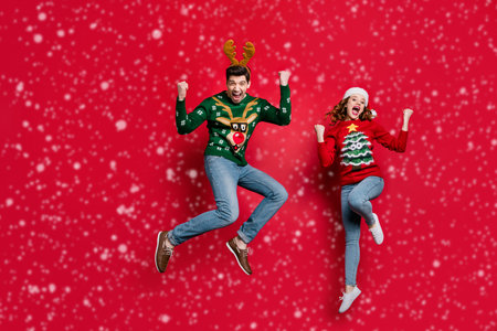 Full length photo of amazed lady and guy jumping excited by x-mas prices wear ugly ornament jumpers and headwear isolated red color backgroundの写真素材
