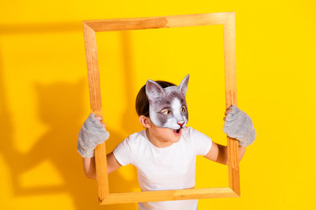 Young girl wearing a grey cat mask holding a wooden frame against a vibrant yellow backgroundの写真素材