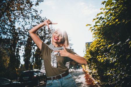 Photo of positive young blonde girl show fingers frame wear khaki outfit tied shoulders sweater walk park sunny spring weather outsideの写真素材