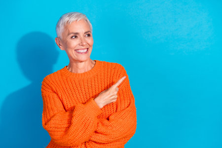 Photo of aged lady look empty space point finger ads wear orange pullover isolated blue color backgroundの写真素材