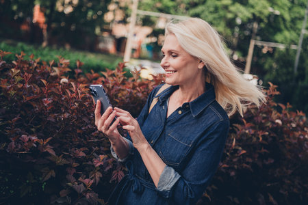 Charming mature woman in denim dress enjoying a sunny day while checking her phone outdoors in a...