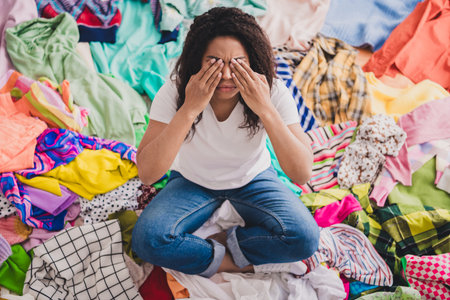 Young woman surrounded by heap of colorful clothes in living room expressing stress