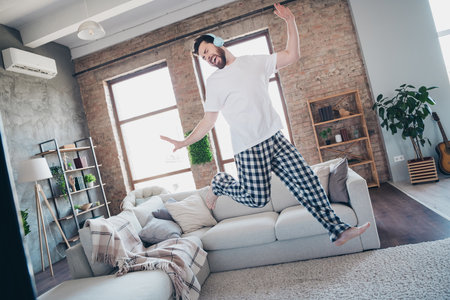 Full length photo of handsome young guy jump dance headphones listen music dressed nightwear wake up spacious house indoors roomの写真素材