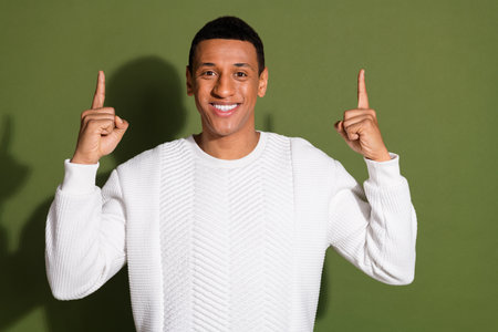 Young man in white sweater pointing upwards with both hands against green backgroundの写真素材