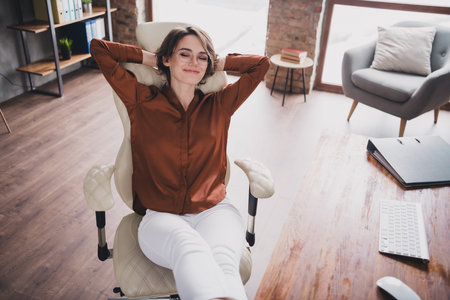 Photo of lovely young lady hands behind head relax sleepy dressed brown satin shirt startup office room interiorの写真素材