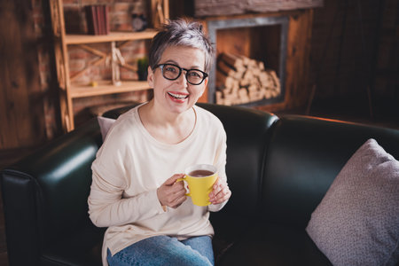 Charming elderly woman with short grey hair and glasses enjoying a cozy moment at home, sipping tea,...